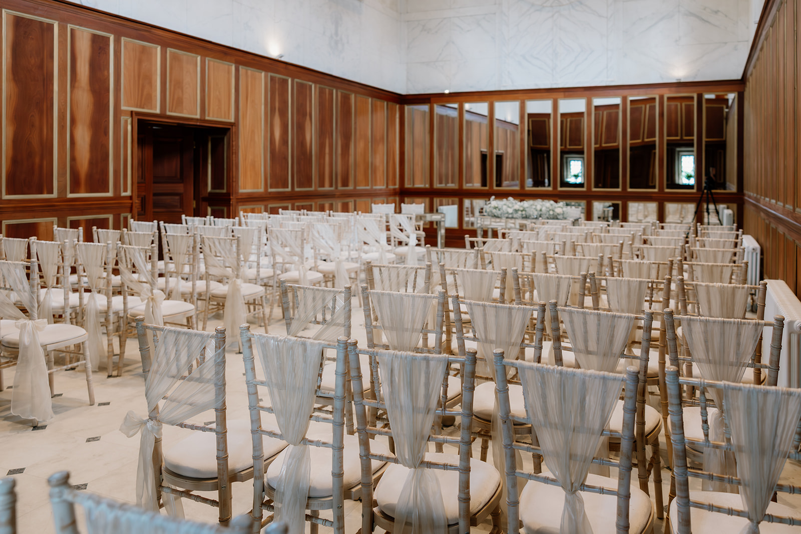 Rows of empty white chairs with sheer fabric draping are arranged in the Chapel, a wood-panelled room with large mirrors, set up for an indoor event or ceremony.