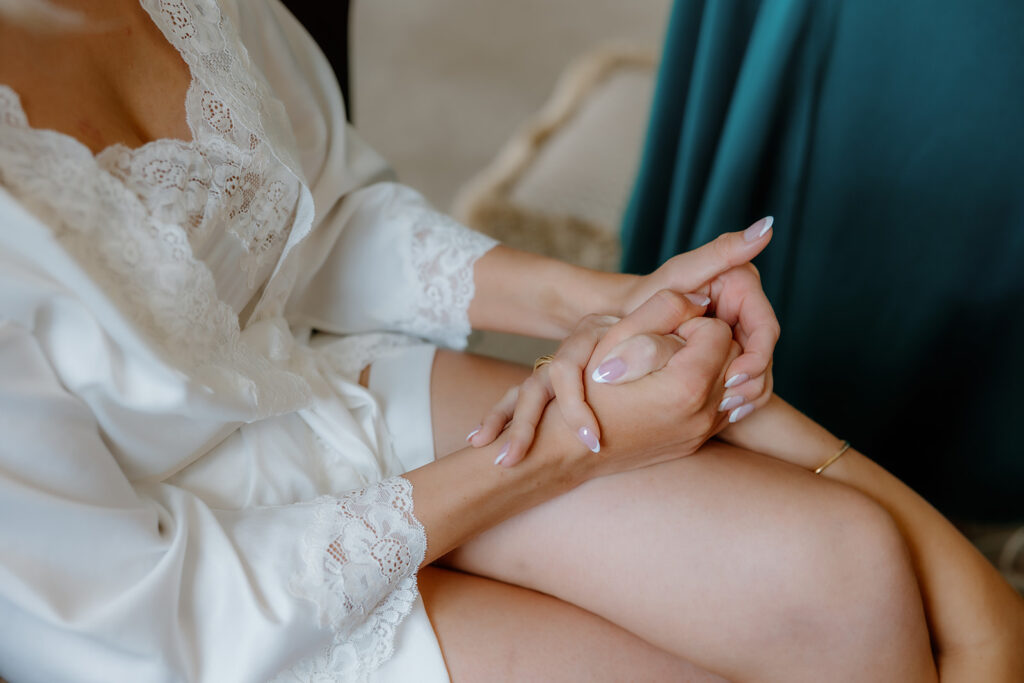 A person in a white lace dressing gown sits with hands clasped on their lap, showing manicured nails and a gold bracelet.