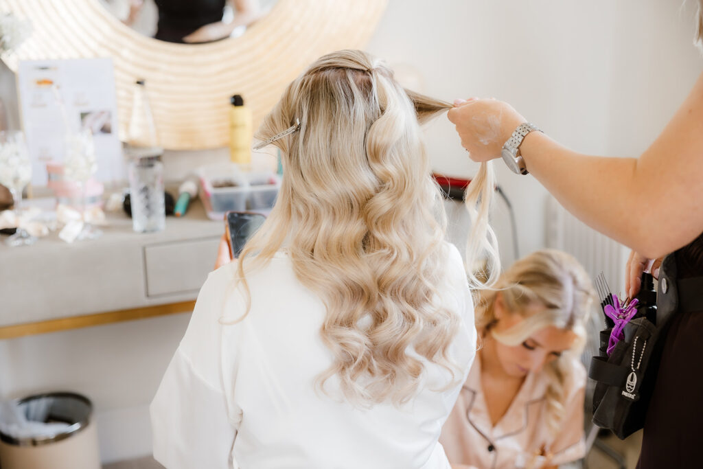 The bride with long, wavy blonde hair sits at a dressing table while another person styles her hair, using clips and a curling tong. A second person is seated on the floor nearby.