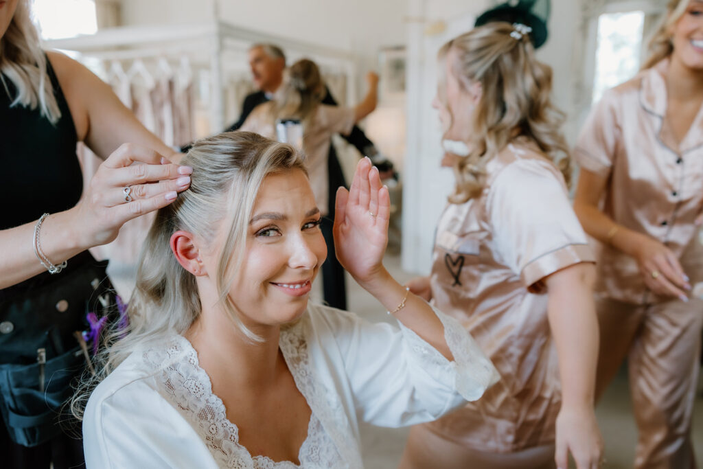 A woman in a dressing gown smiles whilst having her hair styled, surrounded by others in matching satin pyjamas in a bright room.