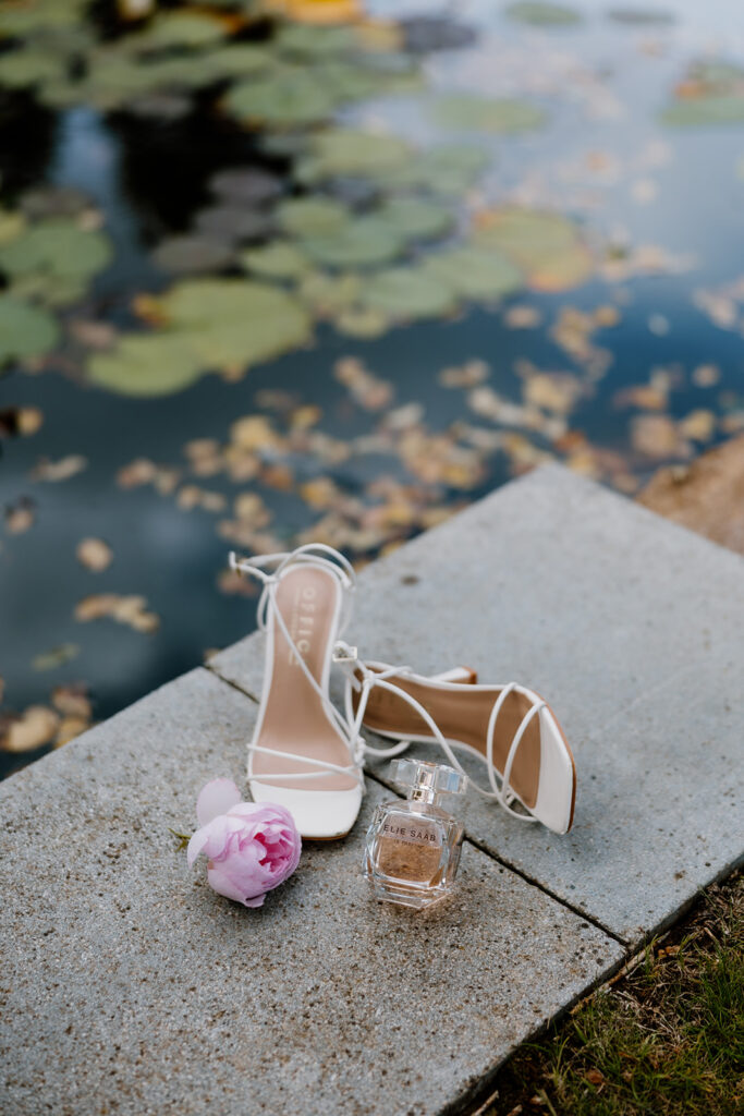 White high-heeled sandals, a pink peony, and a perfume bottle are arranged on a stone surface beside a pond with lily pads and scattered leaves.