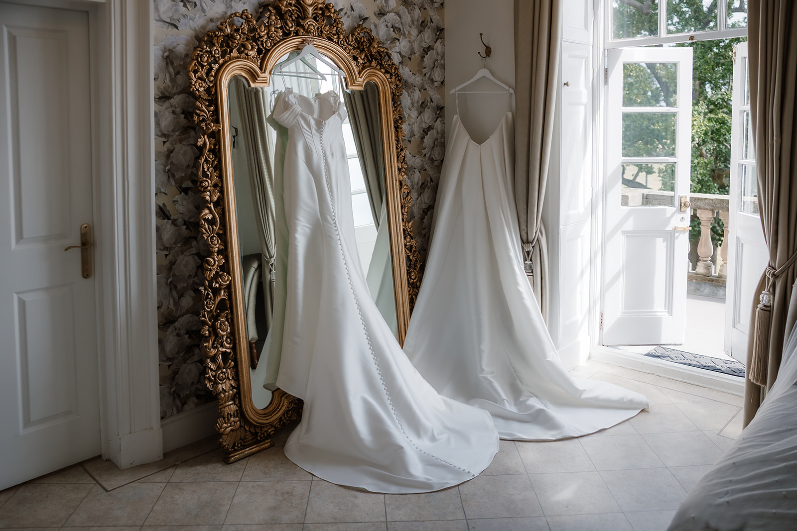 Two elegant white wedding dresses are displayed in a bright room, one wedding dress hanging on a looking glass and the other on a hanger by an open door with a garden view.