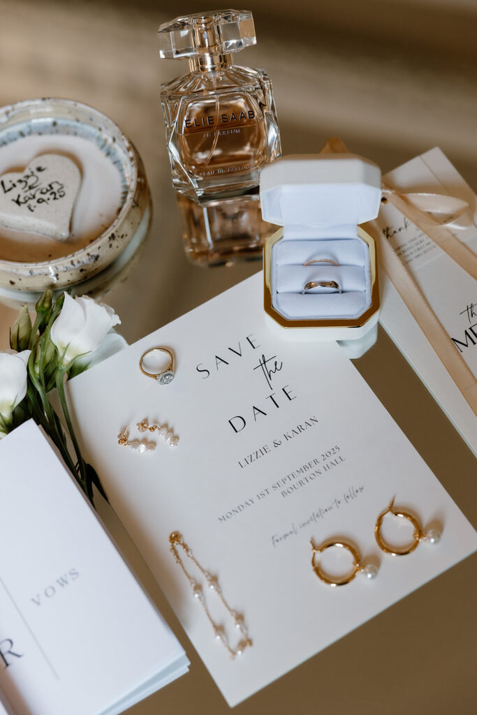 A wedding flat lay featuring a save the date card, rings in a box, perfume, jewellery, white roses, and vow booklets arranged on a reflective surface.