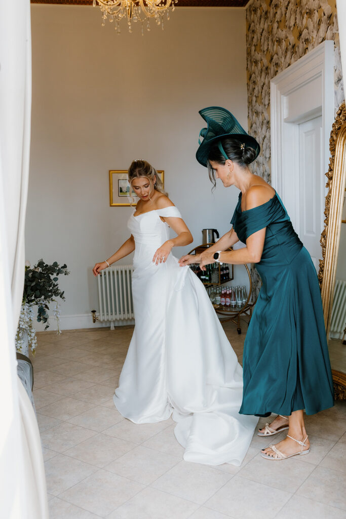 A bride and her mother share a moment as the woman in a teal dress adjusts the train of the bride's white gown in a well-lit room with floral wallpaper and a large mirror.