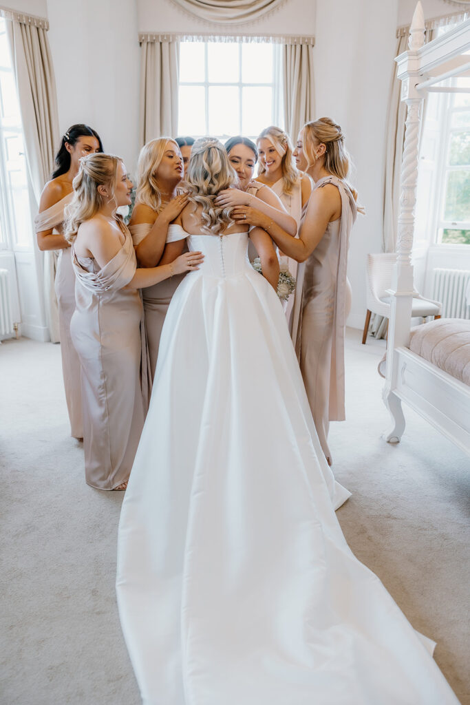 A bride in a white dress is surrounded and embraced by six bridesmaids in light-coloured dresses in a bright, elegant room.