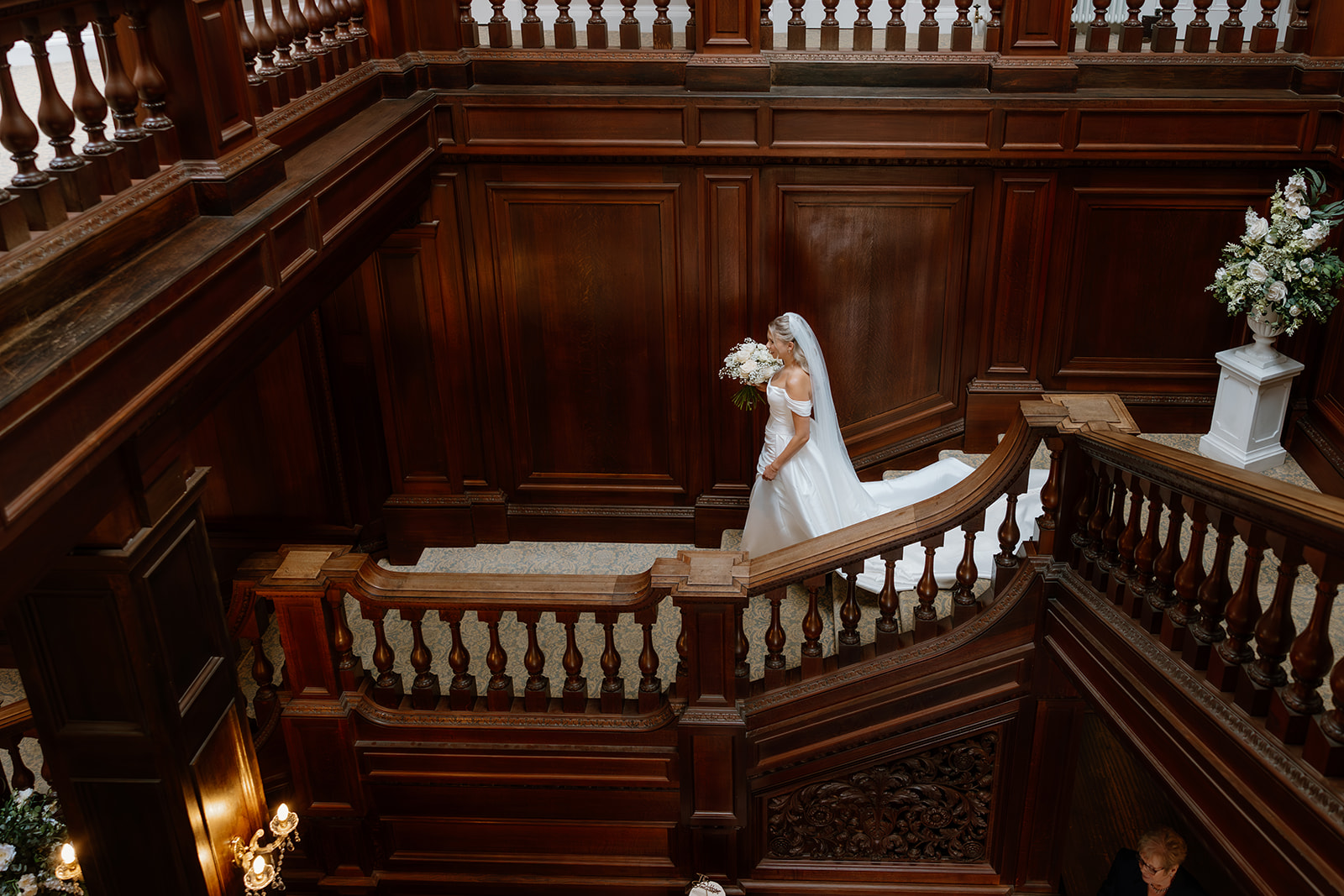 A bride in a white dress and veil holds a bouquet whilst walking down the staircase in an ornately decorated interior.