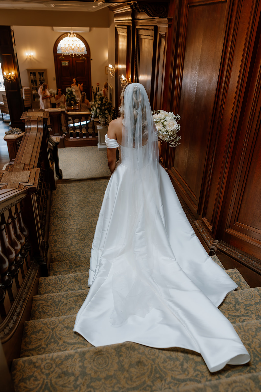 A bride in a white dress and veil descends a carpeted staircase in a wood-panelled interior, holding a bouquet as people gather in the background.
