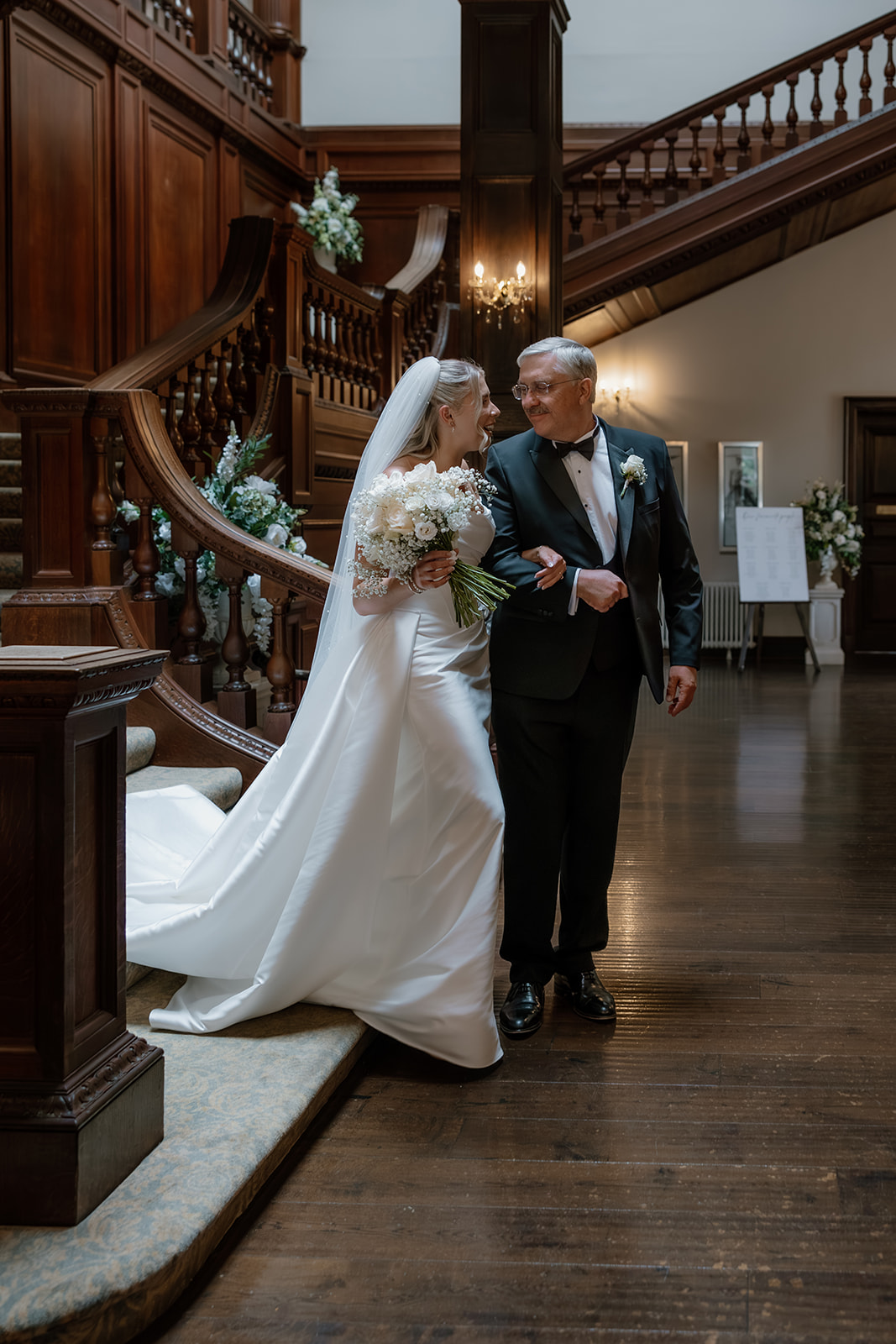 A bride and her father walk arm-in-arm down a wooden staircase in an elegant hall—the bride in a white gown and veil, holding a bunch of flowers, beside her father dressed smartly in a dinner suit.
