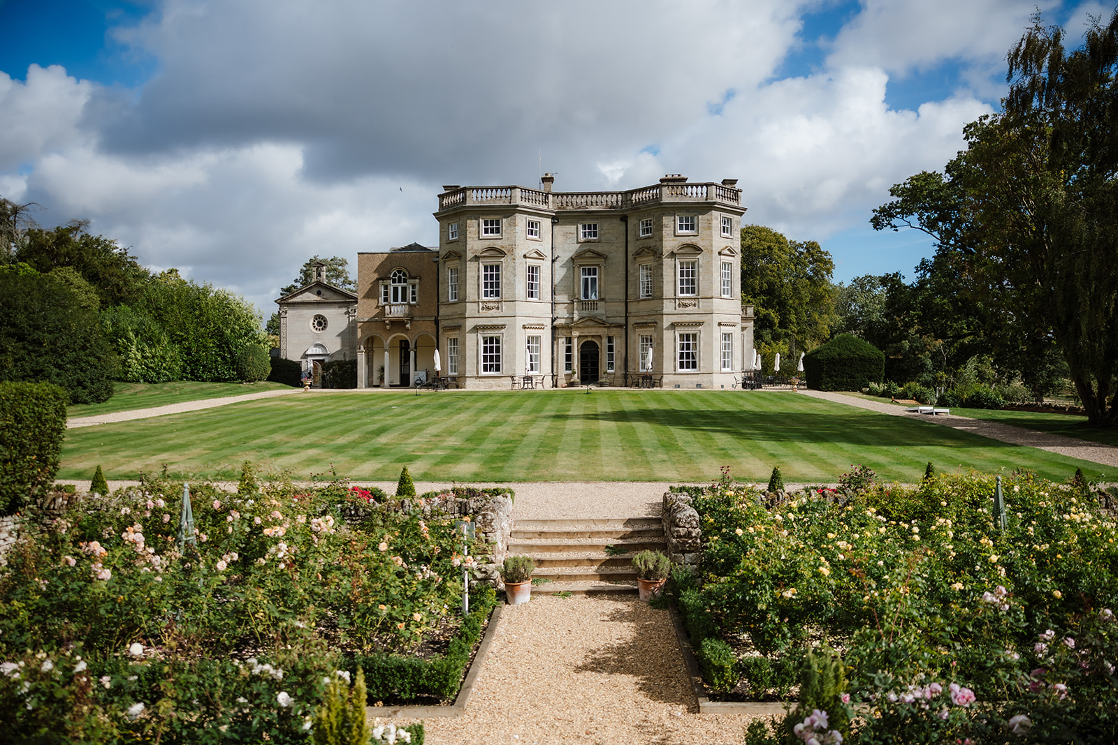 The large historic stone mansion, Bourton Hall, with multiple windows and columns stands behind a manicured lawn and symmetrical garden under a partly cloudy sky.