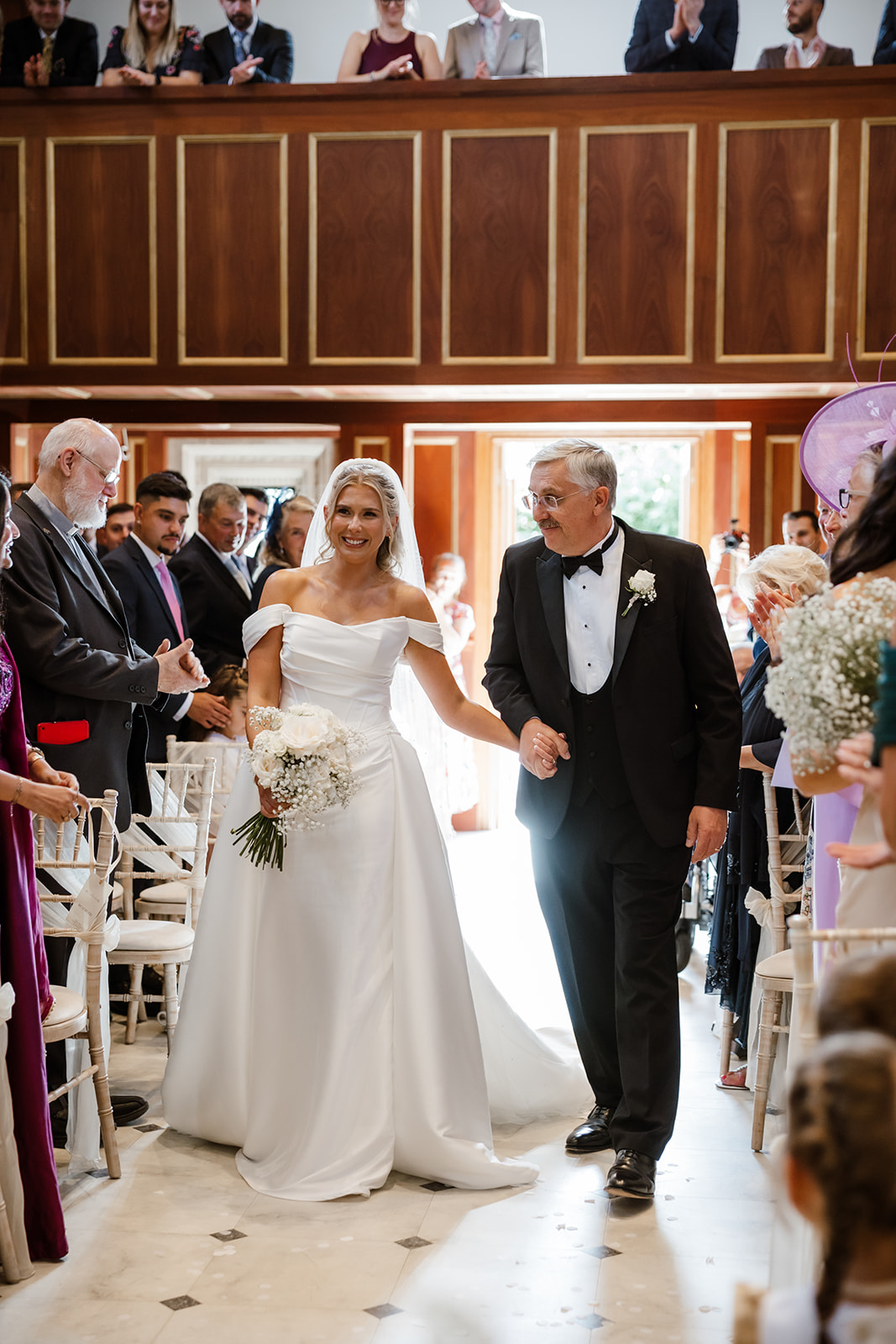 A bride in a white dress walks down the aisle with an older man in a dinner jacket, surrounded by seated and standing guests in a wood-panelled venue.