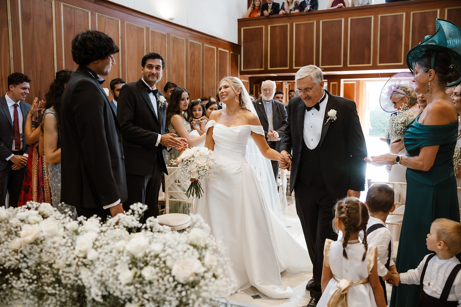 Bride in white dress walks down the aisle with an older man; guests watch and smile in a wood-panelled room decorated with white flowers.