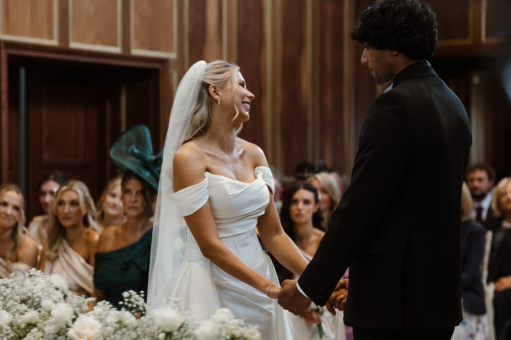 A bride and groom stand facing each other, holding hands during their wedding ceremony, with guests seated and watching in the background.