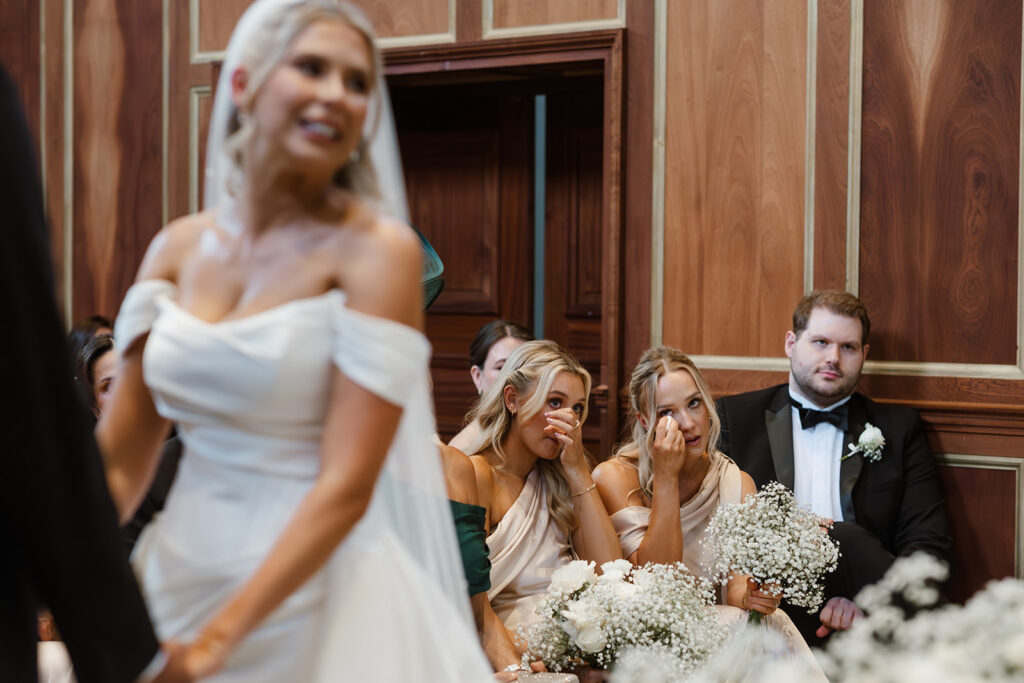 Two emotional bridesmaids wipe away tears whilst sitting and holding bouquets during a wedding ceremony; a bride in a white dress is in the foreground.