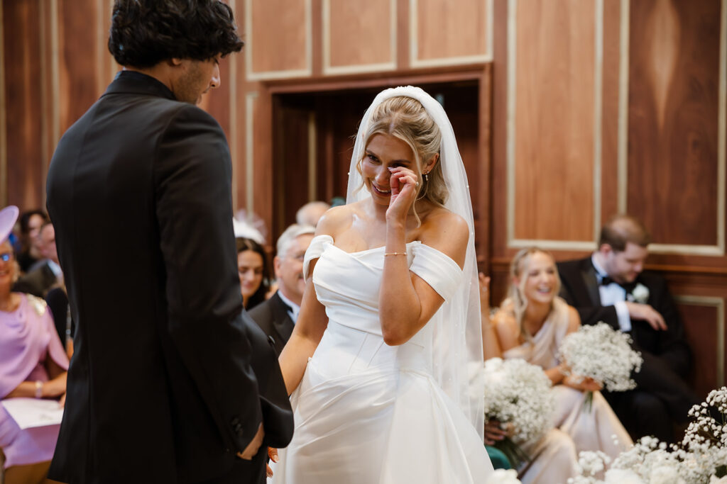 A bride in a white dress wipes away a tear whilst standing next to a groom in a black suit during a wedding ceremony, with seated guests watching in the background.