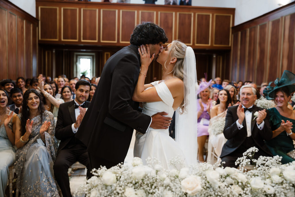 A bride and groom kiss at the altar in a wood-panelled room, surrounded by applauding guests. White flowers decorate the foreground.