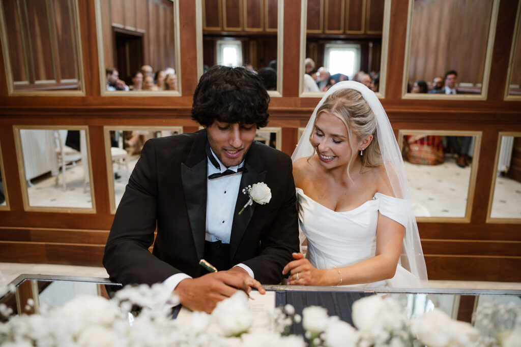 A groom in a dinner suit signs a document while a bride in a white dress and veil sits beside him, both smiling, in a wood-panelled room with people in the background.