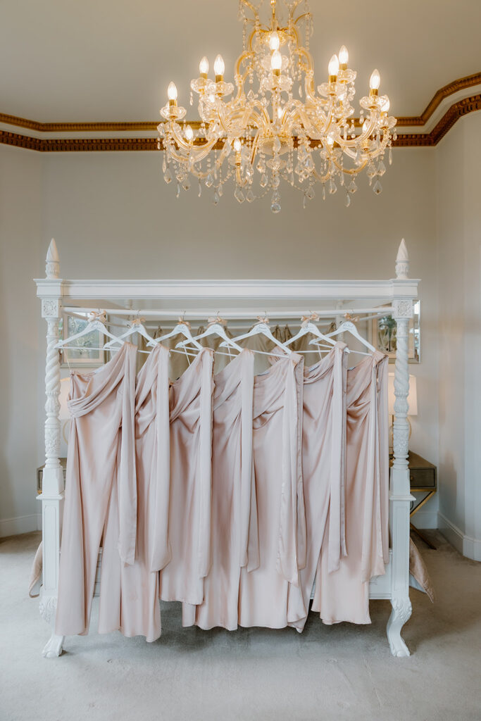 Six blush-coloured dresses on hangers are arranged on a white canopy bed frame beneath an ornate chandelier in a bright room.