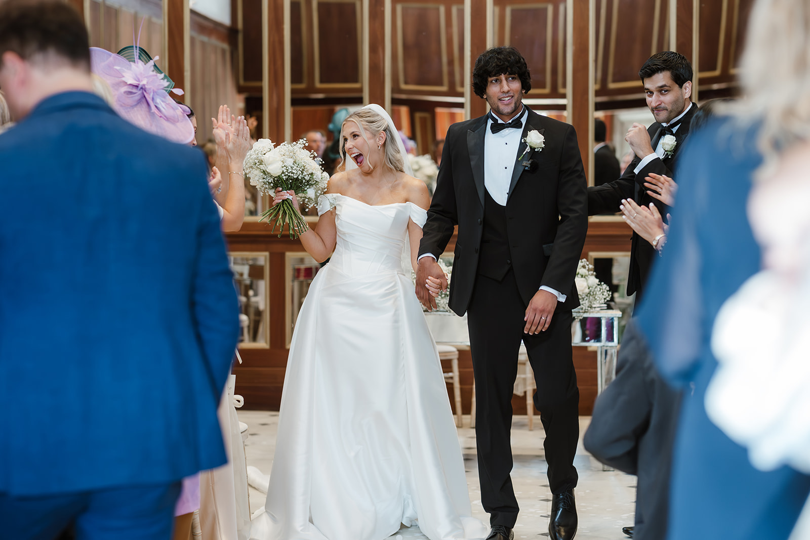 A bride and groom, both smiling, walk hand in hand down an aisle as guests clap and celebrate around them at an indoor wedding ceremony.