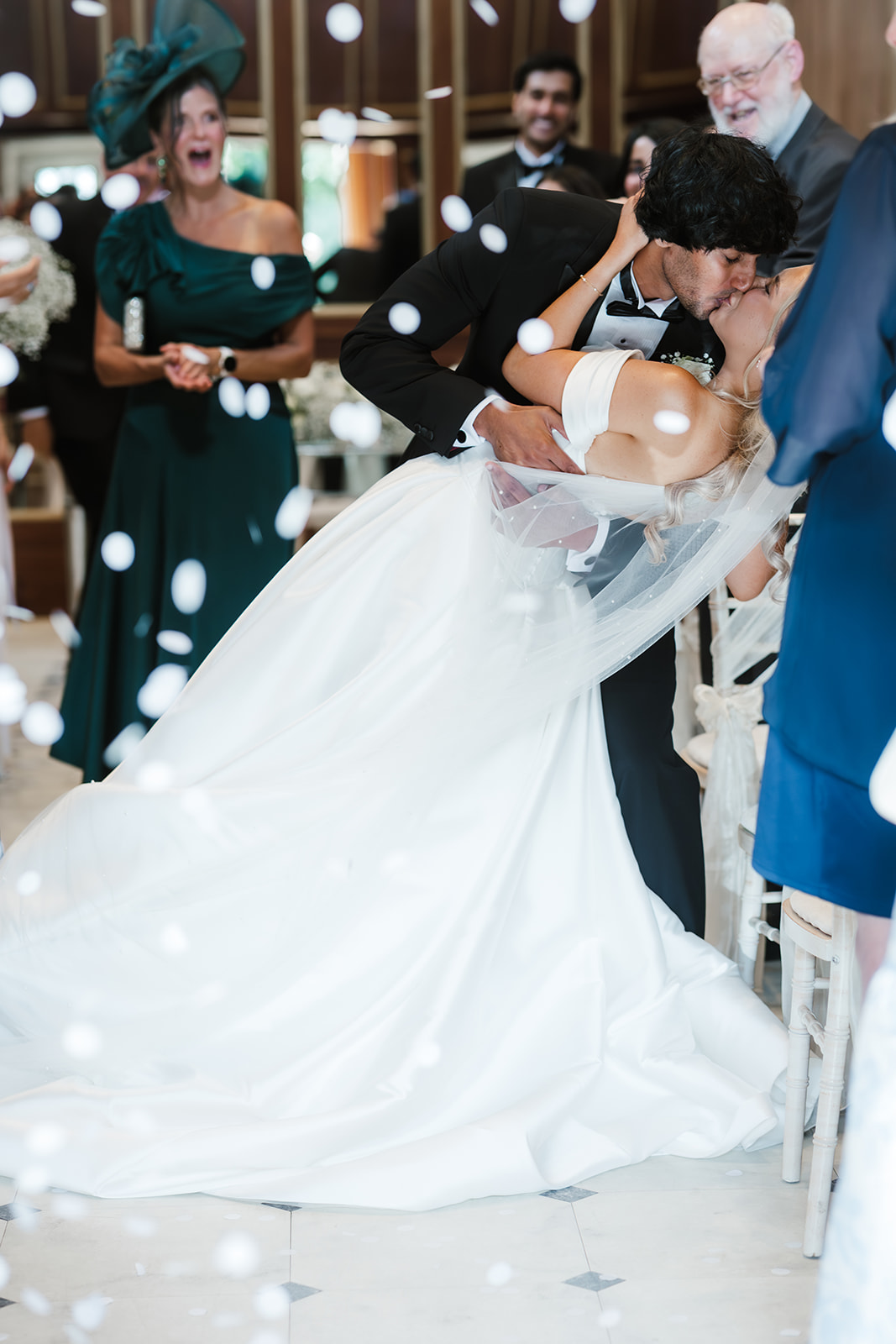 A bride and groom kiss during their wedding ceremony as guests watch and white confetti floats in the air.