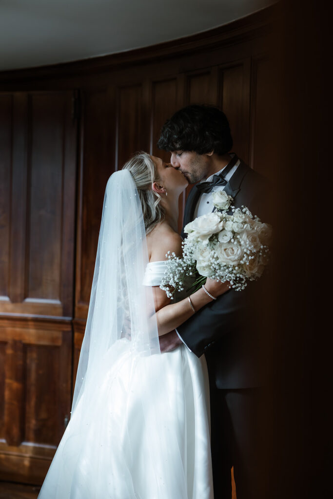A bride and groom embrace indoors, with the bride holding a bouquet of white flowers and wearing a veil, both dressed in formal wedding attire.