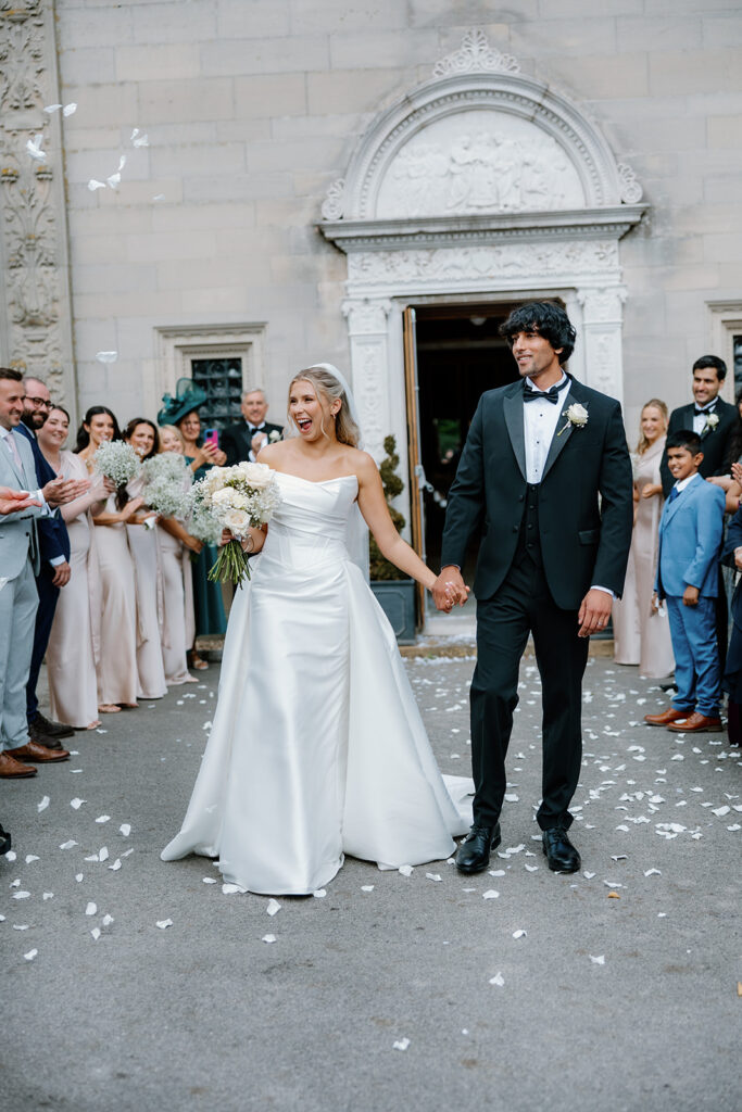 A bride and groom hold hands outside Bourton Hall, surrounded by smiling wedding guests. The bride holds a bouquet, and confetti petals are scattered on the ground.