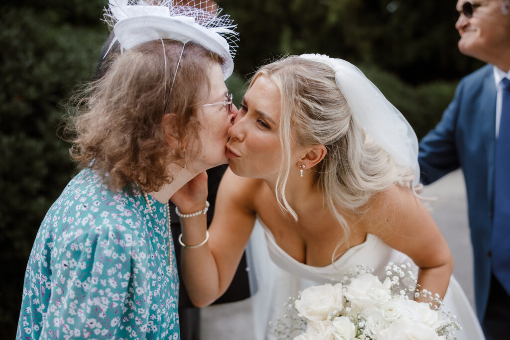 A bride in a white dress and veil kisses an elderly woman on the cheek outdoors, holding a bouquet of white flowers.