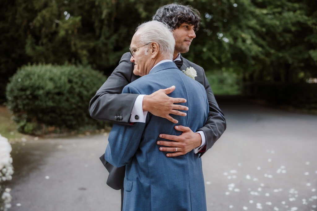 A younger man in a suit embraces an older man in a blue jacket on a tree-lined path, with flower petals scattered on the ground.