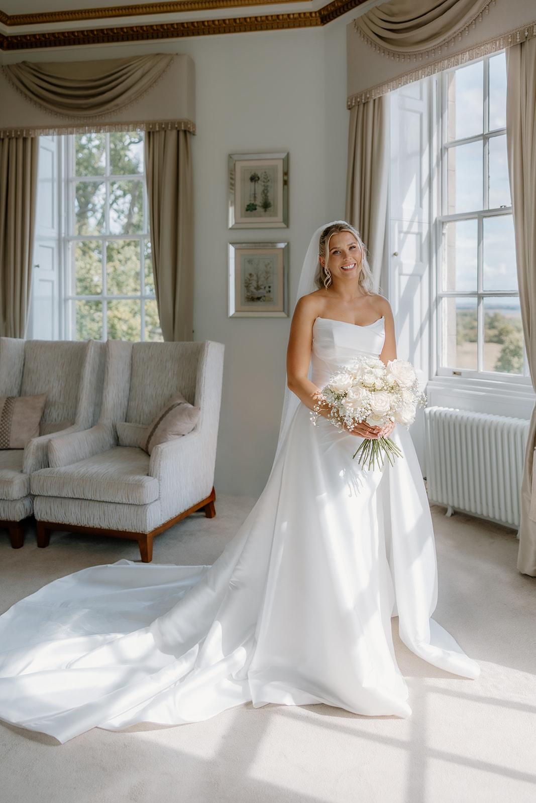 A bride in a white wedding dress stands indoors by the grand windows of Bourton Hall, holding a bouquet of white flowers as sunlight streams into the elegant room.