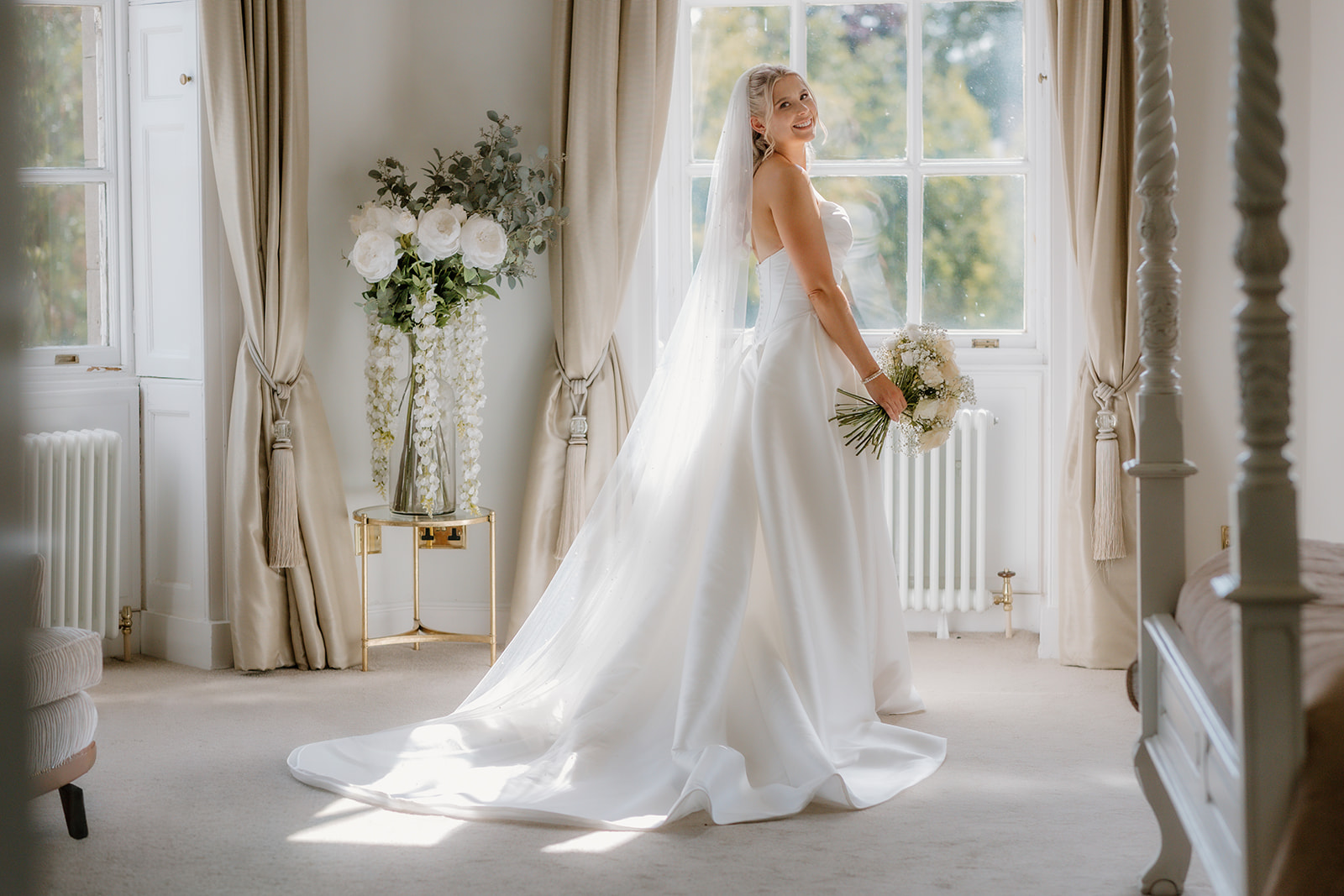 A bride in a white wedding dress and veil stands in a bright, elegant room at Bourton Hall, holding a bouquet, sunlight streaming through the window behind her—perfectly captured by her wedding photographer.