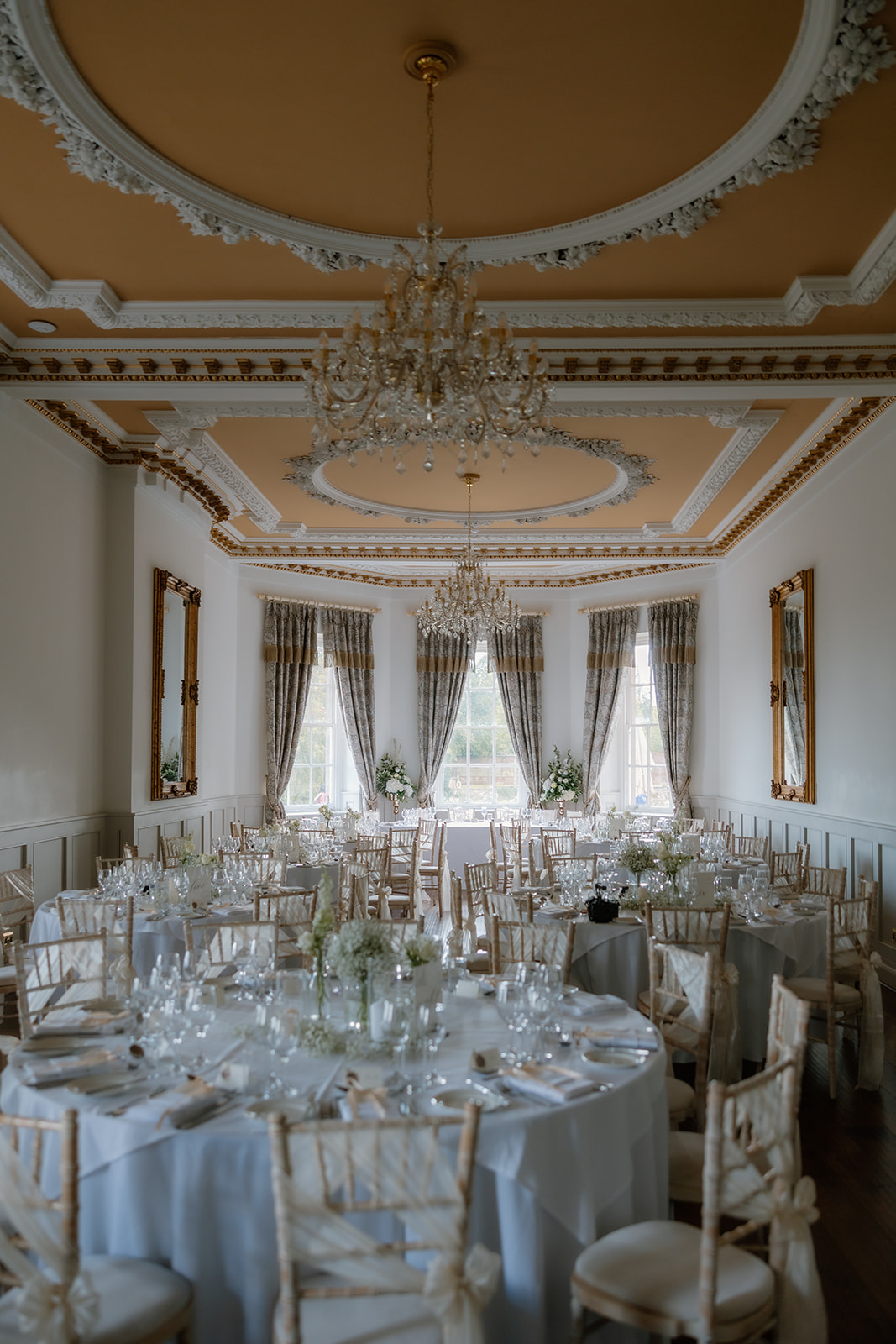 Elegant banqueting hall with round tables set for wedding breakfast, featuring gold-trimmed ceiling, chandeliers, large mirrors, and tall windows with draped curtains.