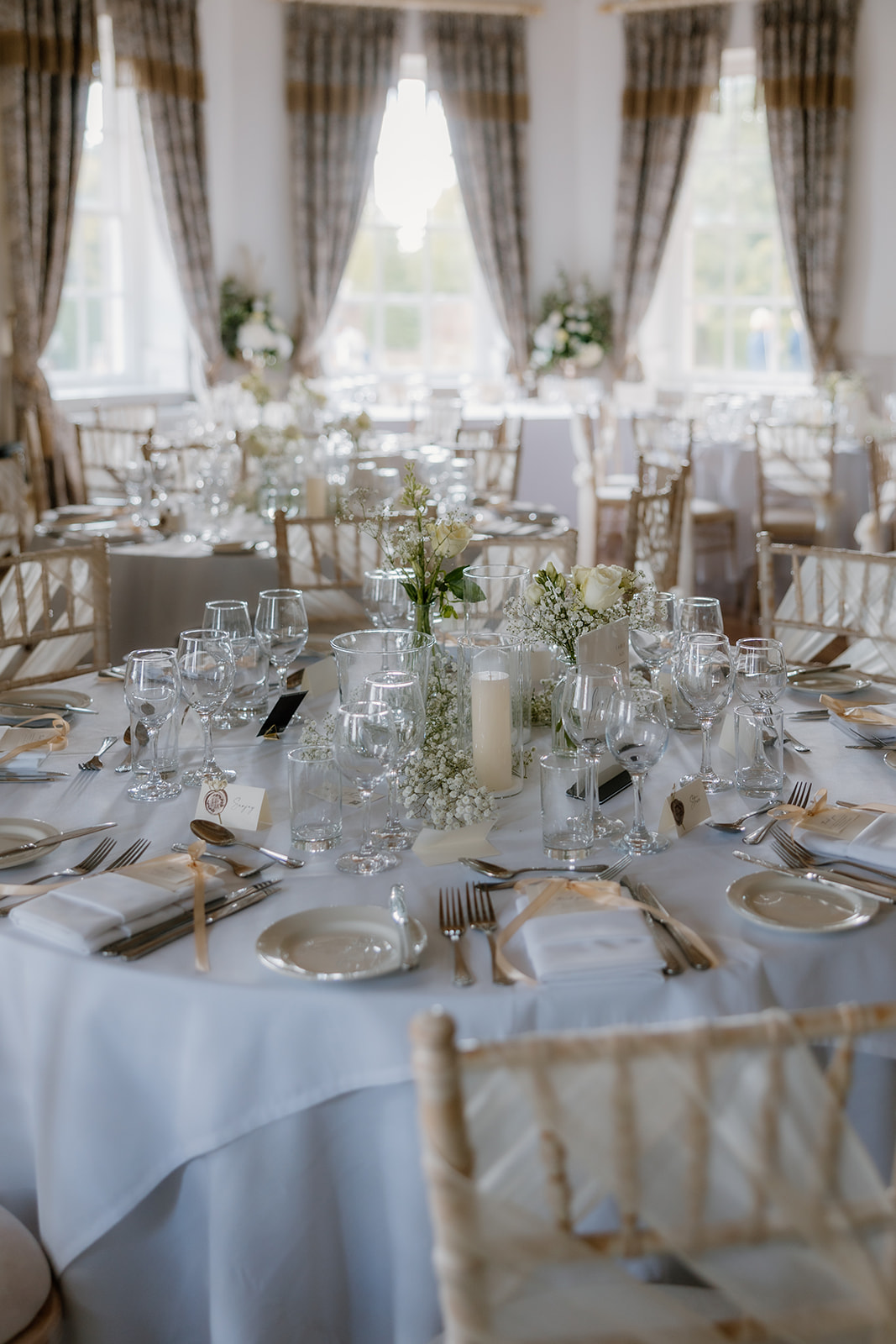A round banquet table set for a wedding breakfast, featuring glassware, plates, cutlery, a white tablecloth, candles, and floral centrepieces in a bright, elegant room.