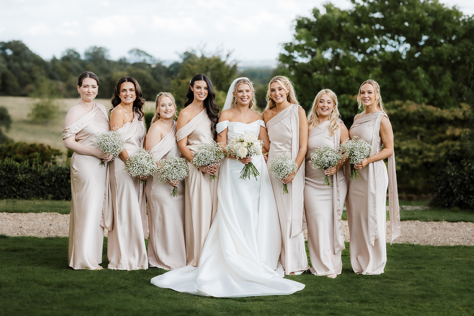 A bride in a white dress stands outdoors with seven bridesmaids in matching light pink dresses, all holding bouquets of gypsophila.