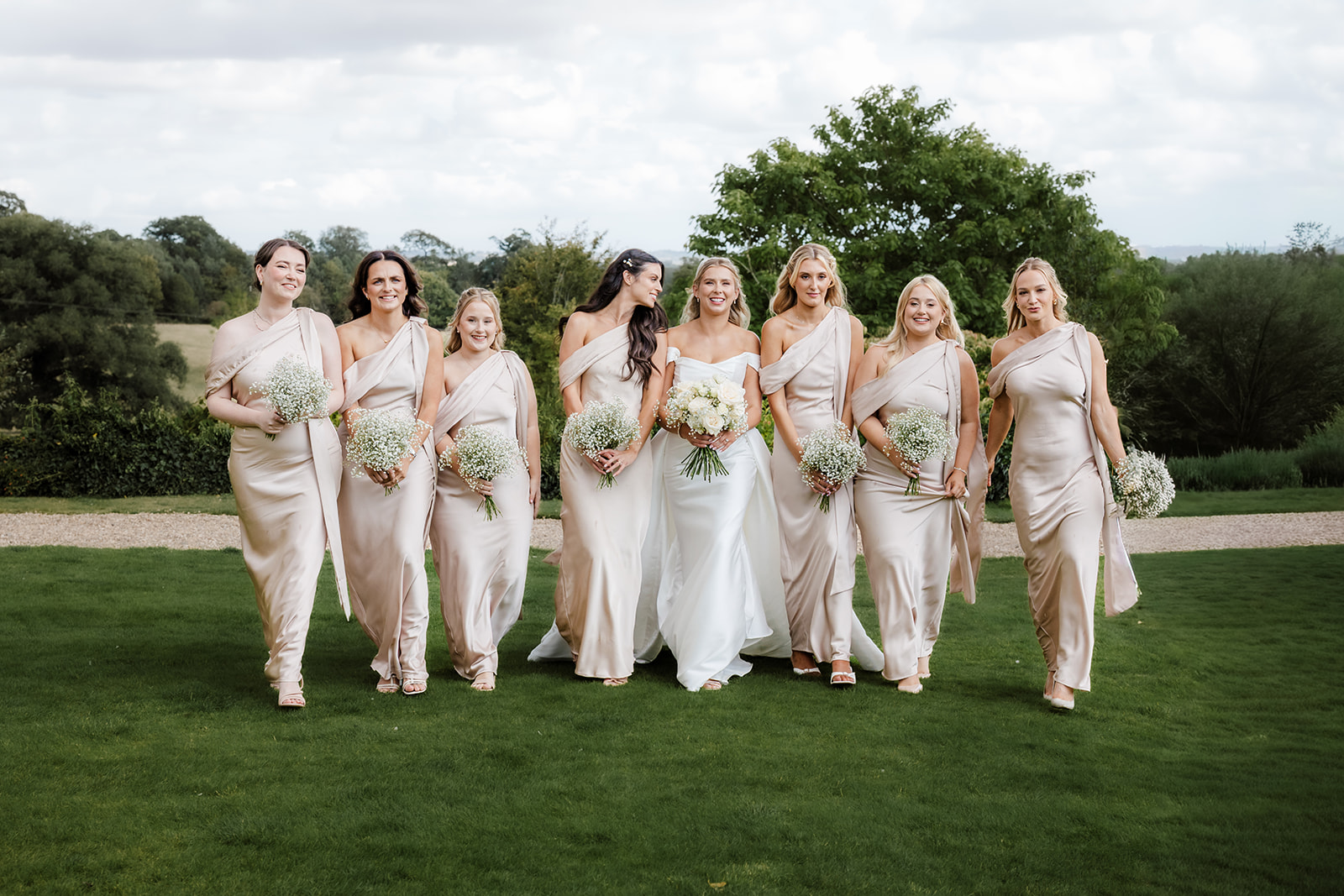 A bride in a white dress stands with eight bridesmaids in light-coloured gowns, all holding bouquets, walking together on a lawn with trees in the background.