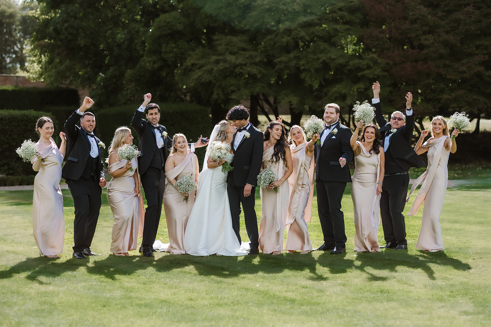 A bride and groom kiss whilst standing with their wedding party, who cheer and hold flowers, on a lawn with trees in the background.