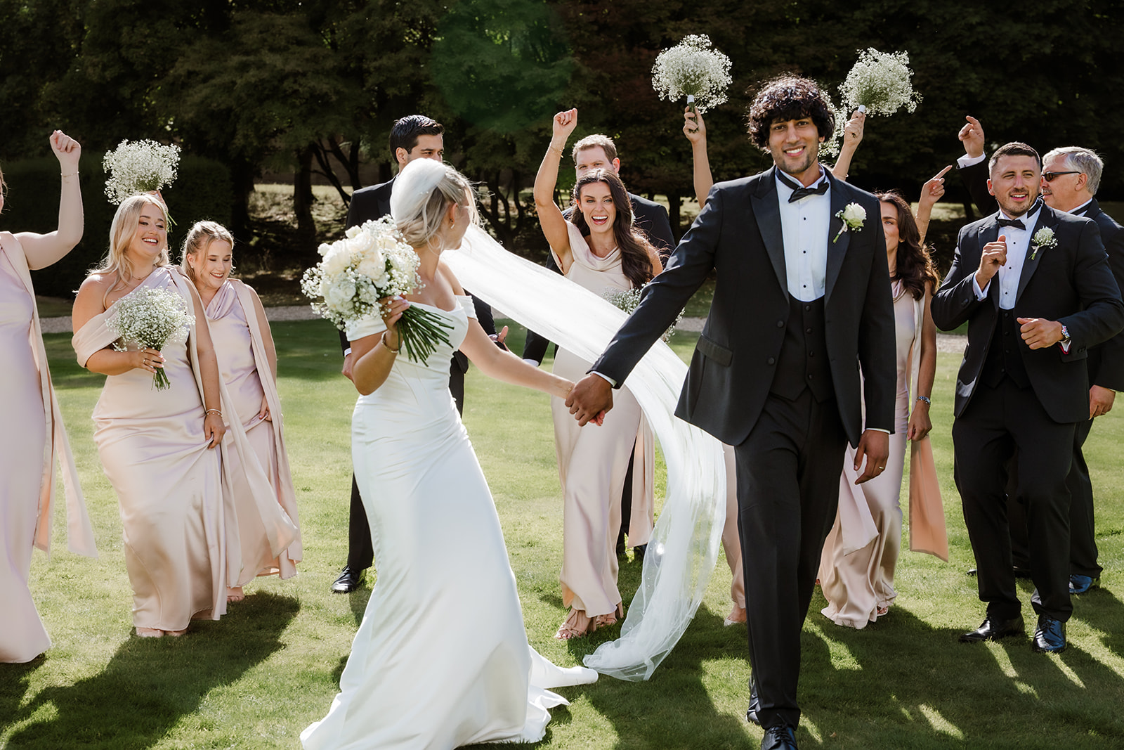 A bride and groom walk hand in hand outdoors, smiling, surrounded by cheering bridesmaids in pale dresses and groomsmen in dark suits holding bouquets at The Bourton Hall Wedding.