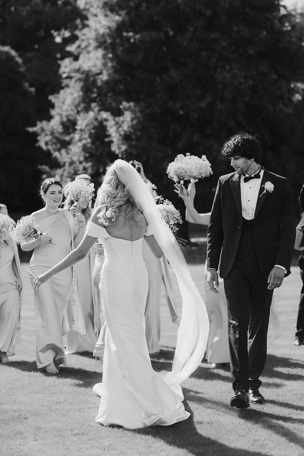 A bride in a long gown and veil walks on grass with bridesmaids holding posies, while the groom in a suit looks at her.