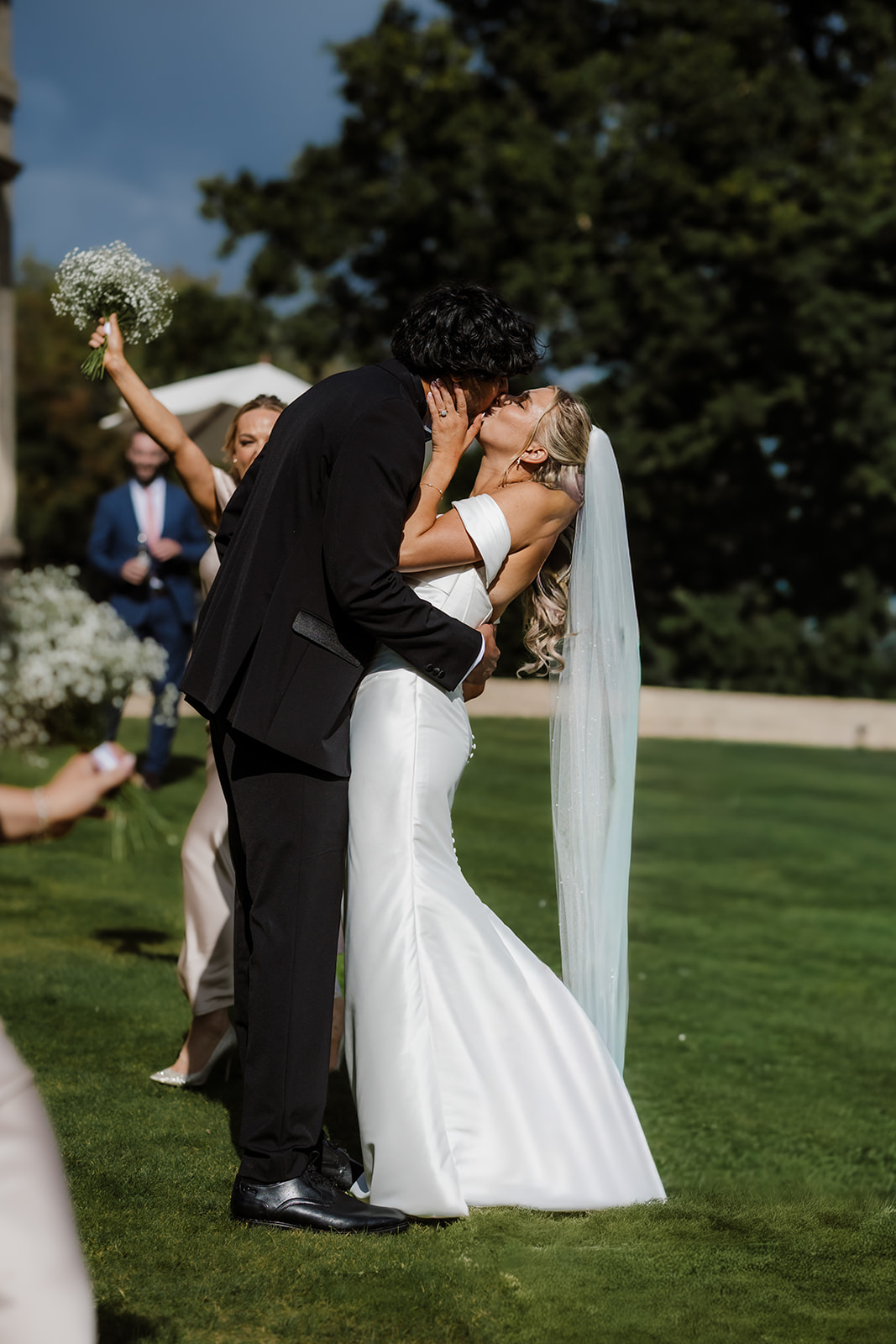 A bride and groom kiss outdoors on a grassy lawn, with the bride in a white dress and veil and the groom in a black suit. Bridesmaids are visible in the background.