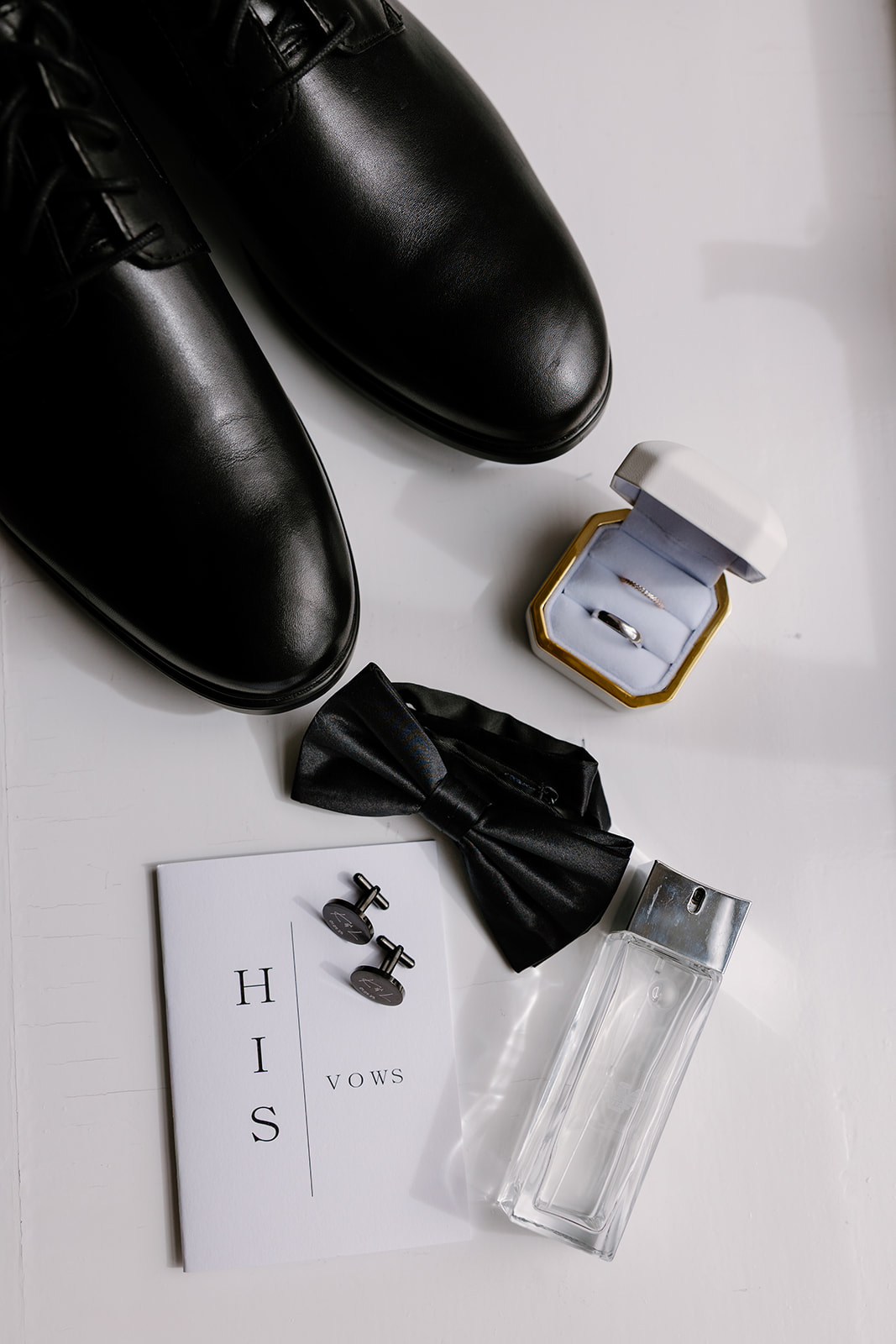 Black dress shoes, a bow tie, a pair of cufflinks, a bottle of aftershave, an open ring box with a ring, and a "His Vows" booklet arranged on a white surface.
