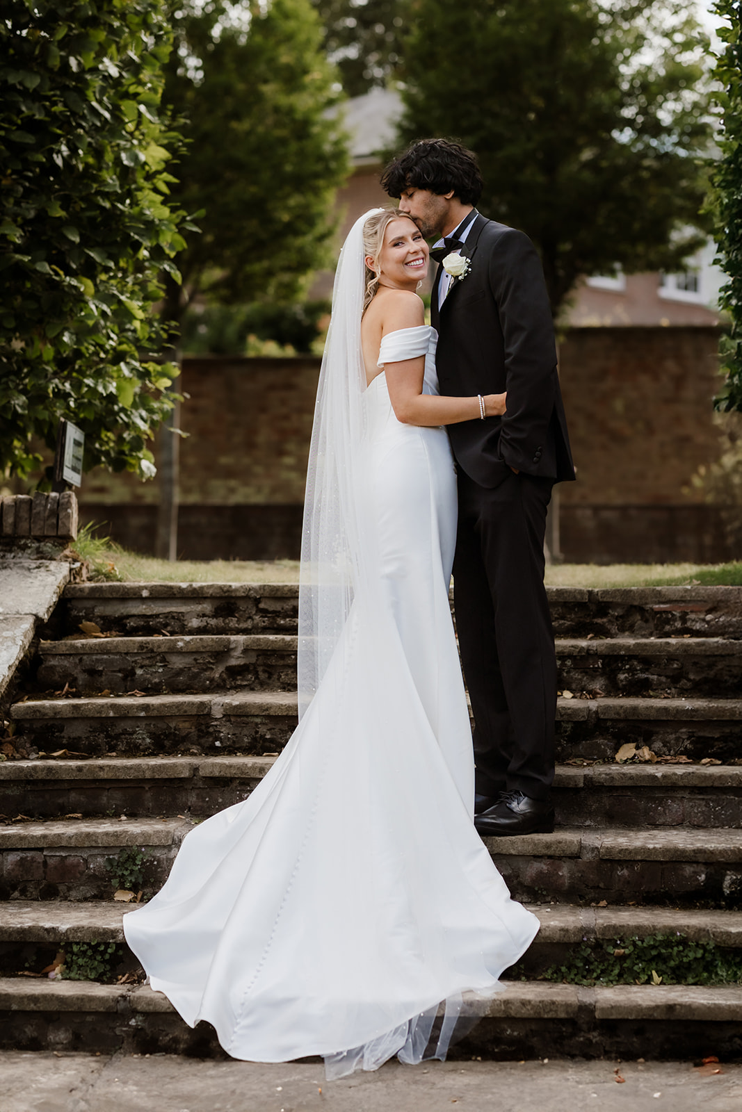 A bride in a white off-shoulder gown and veil stands on stone steps, smiling, as the groom in a black suit kisses her on the head. Outdoors, greenery and a brick wall are in the background, Bourton Hall Wedding Photographer.