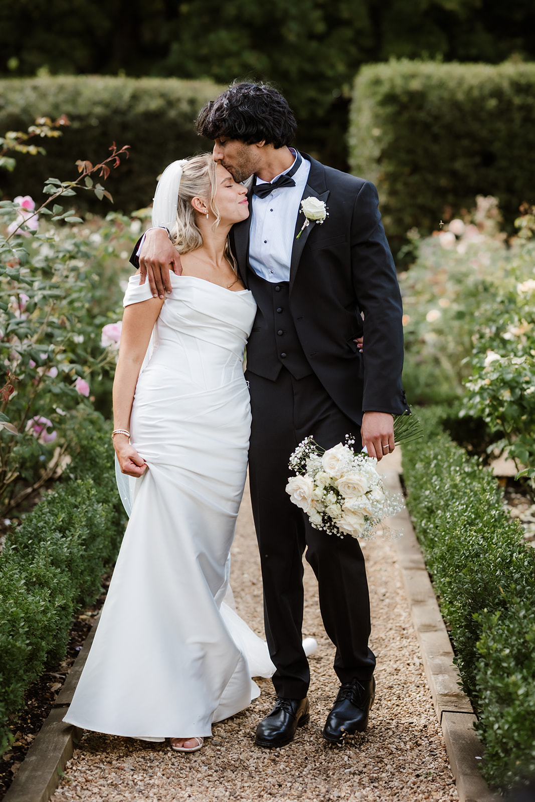 A bride and groom in formal wedding attire stand close together on a garden path, with the groom holding a bouquet and embracing the bride.