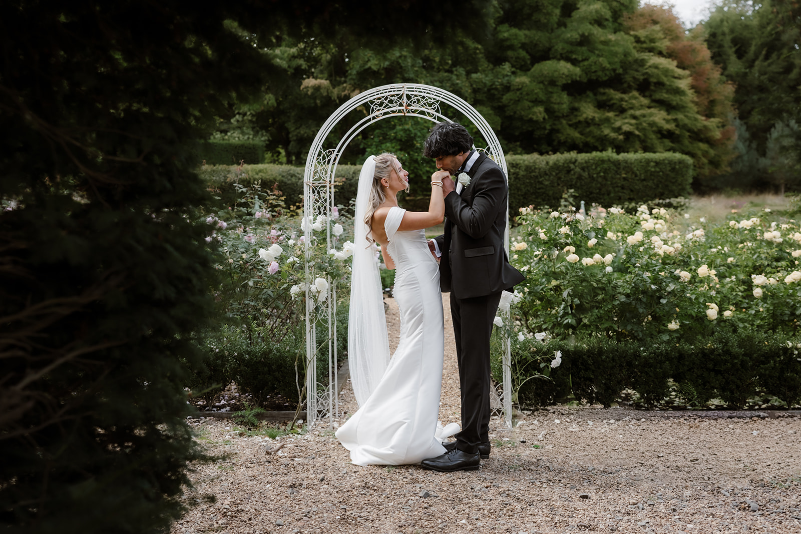 A bride and groom stand under a white arch in a garden; the groom kisses the bride’s hand whilst both smile at each other. Bourton Hall Wedding Photography. 