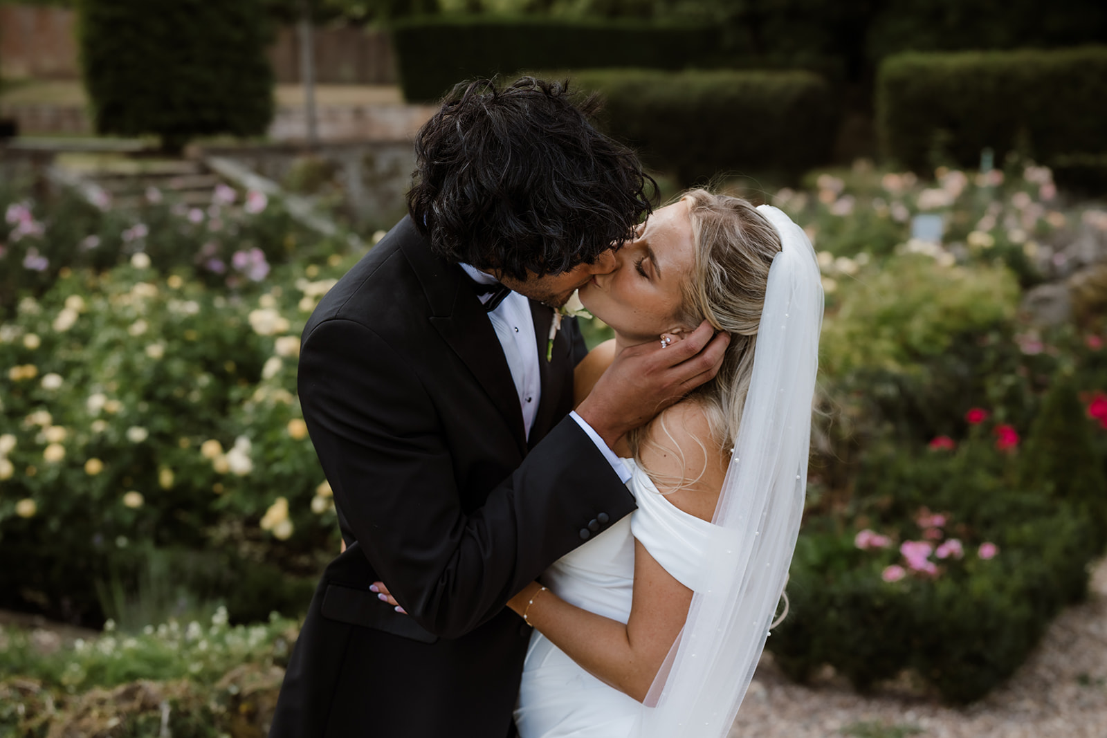 A bride and groom in formal attire share a kiss in a garden, surrounded by blooming flowers and greenery. Bourton Hall Wedding Photographer