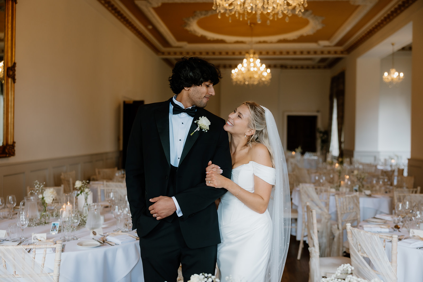 A bride and groom stand together in an elegantly decorated reception hall, looking at each other and smiling in the wedding breakfast room at Bourton Hall.