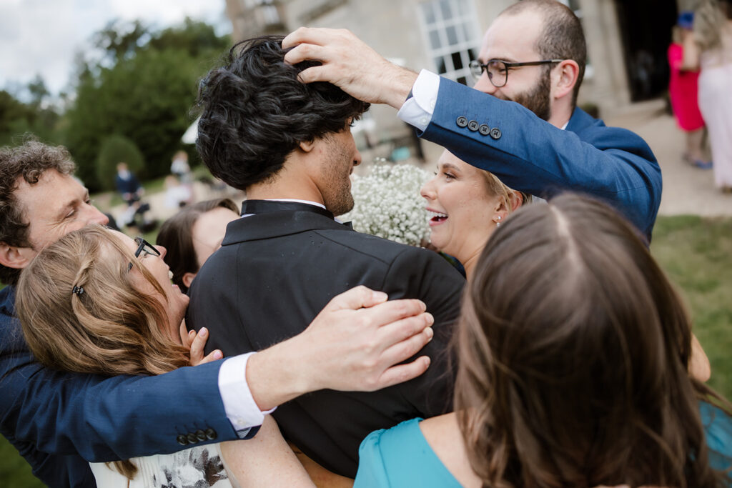 A group of people in formal attire gather closely, hugging and laughing outdoors, with one person playfully adjusting another’s wig or hairpiece.