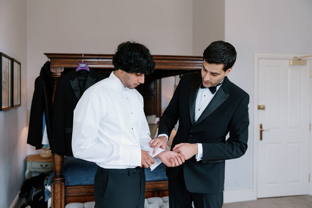 A groom and his brother, both in tuxedos, stand in a bedroom as one helps the other fasten his shirt cuffs. A suit jacket hangs in the background.