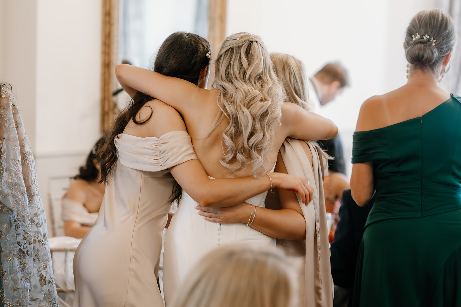 Three women in formal dresses stand with their arms round each other, facing away from the camera at an indoor event.