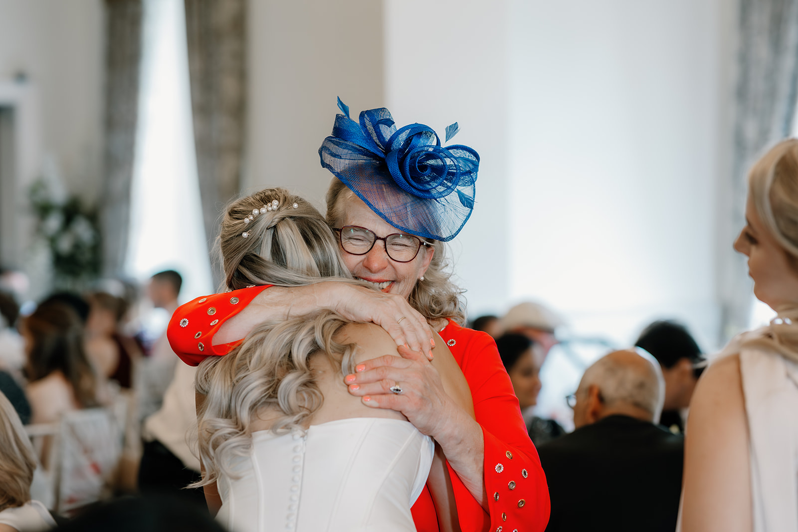 An older woman in a blue hat and red jacket hugs a bride in a white dress at an indoor event, with people seated in the background.