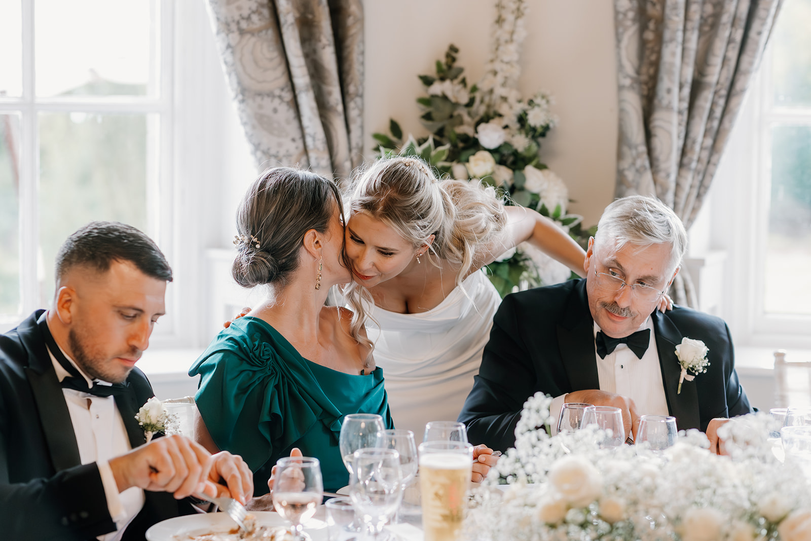 A woman in a white dress leans over to kiss a seated woman on the cheek at a formal event, while two men in dinner jackets sit nearby at a table set with food and flowers.