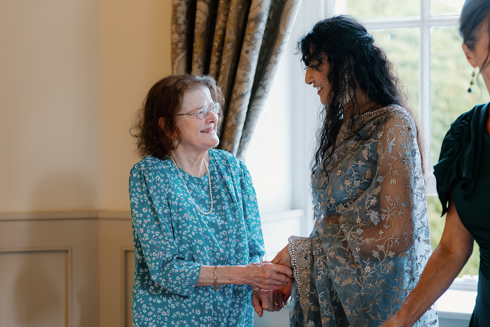 An older woman in a blue patterned dress smiles and holds hands with a younger woman in an embroidered sari, standing indoors near a window.